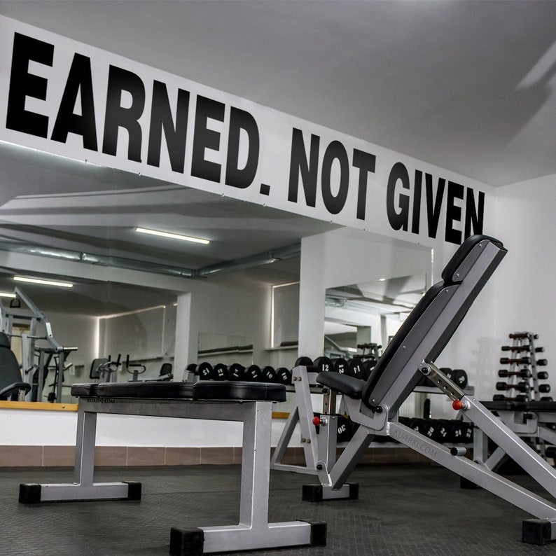 May include: A black and white sign with the text "Earned. Not Given." hanging above a weight bench in a gym. The bench is made of metal and has a black padded seat. The gym is empty and has a white ceiling and walls.