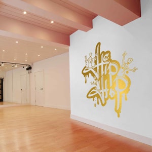 K&ouml;nnte beinhalten: Eine wei&szlig;e Wand mit einem goldenen Spray-Design, auf dem "The Hip Hop" steht, mit einer Cartoon-Figur, die Spraydosen h&auml;lt.