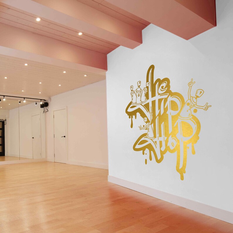 K&ouml;nnte beinhalten: Eine wei&szlig;e Wand mit einem goldenen Spray-Design, auf dem "The Hip Hop" steht, mit einer Cartoon-Figur, die Spraydosen h&auml;lt.