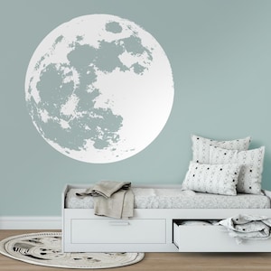 Può includere: Un adesivo da parete a forma di luna bianca con una superficie testurizzata, applicato su una parete blu chiaro in una stanza con un divano letto bianco e un tappeto a motivi.