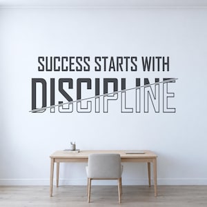 Può includere: Un adesivo da parete con il testo grigio scuro SUCCESS STARTS WITH DISCIPLINE, in cui la parola DISCIPLINE è visualizzata in un carattere grande e in grassetto, attraversata da una sottile linea diagonale, sopra una scrivania e sedia in legno.