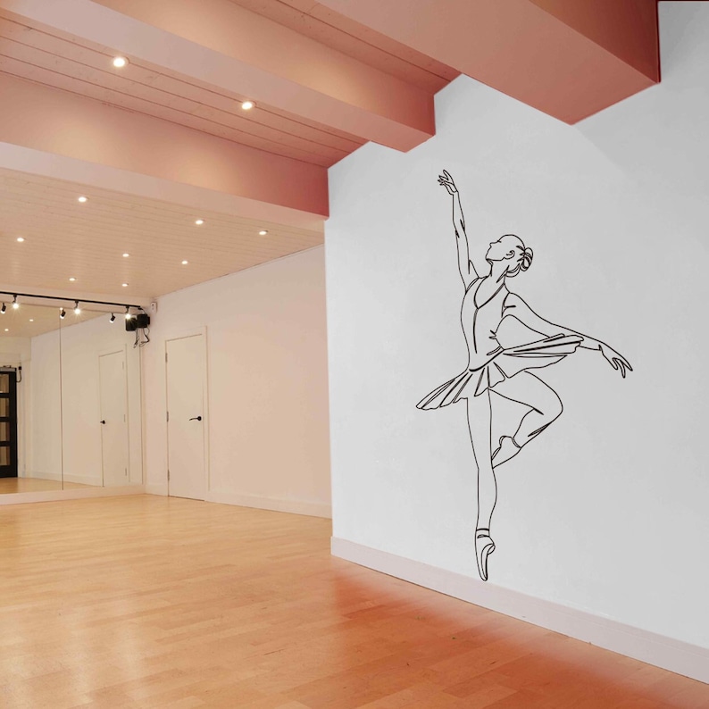 May include: A black line drawing of a ballerina in a white tutu, on a white wall in a dance studio. The studio has a light wood floor and a pink ceiling with recessed lighting.
