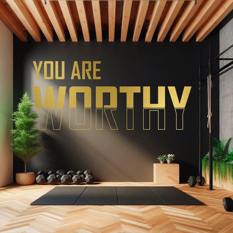 May include: A motivational quote, "You Are Worthy", in gold lettering on a black wall in a gym setting. The quote is displayed on a black wall with a wooden floor and a yoga mat.
