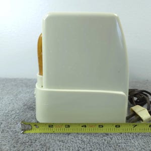 May include: An off-white, vintage electric toaster with a wooden handle. The toaster is rectangular with rounded edges and a power cord. A yellow measuring tape is placed in front of the toaster, showing a length of approximately 15 cm.
