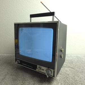 Puede incluir: Un televisor Sony Solid State vintage con un marco negro y plateado, un asa y una antena. La pantalla muestra un tono azul. El televisor tiene varias perillas y botones. El número de modelo es visible.