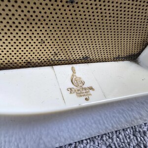 May include: Close-up of a vintage radio or television. The image shows a cream-coloured, curved plastic frame with a gold-coloured, perforated metal speaker cover. The Emerson Radio and Television logo is visible.