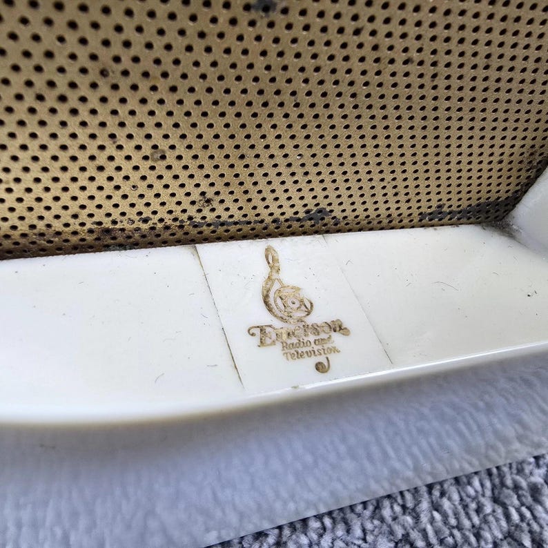May include: Close-up of a vintage radio or television. The image shows a cream-coloured, curved plastic frame with a gold-coloured, perforated metal speaker cover. The Emerson Radio and Television logo is visible.