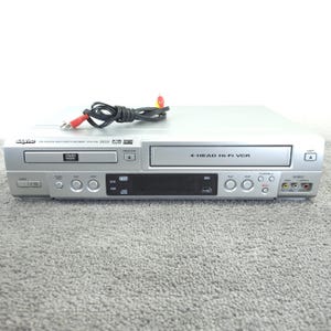 May include: A silver Sanyo DVD player and video cassette recorder combo unit featuring a 4-head Hi-Fi VCR, a DVD disc tray, a central digital display, multiple control buttons, and front-facing AV input ports, accompanied by a black power cable.