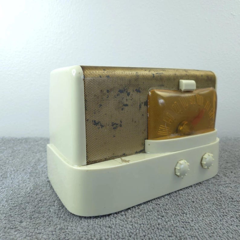 May include: A vintage cream radio with a gold mesh speaker cover and a clear amber dial. The radio has two white knobs and a small white button. The radio is a retro style.