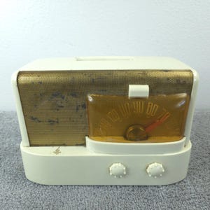 May include: A vintage cream-coloured radio with a gold speaker grille and a clear amber dial. The radio features two white knobs and a red needle on the dial. The radio is a retro design.