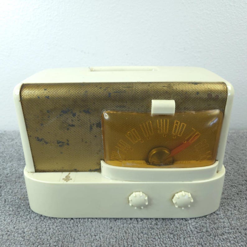 May include: A vintage cream-coloured radio with a gold speaker grille and a clear amber dial. The radio features two white knobs and a red needle on the dial. The radio is a retro design.