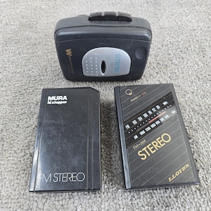 May include: A black portable cassette player with a silver accent and the word "STEREO" printed on it. Two black cassette players are also in the image, one with "MURA hi stepper FM STEREO" printed on it, and the other with "LLOYD'S STEREO FM-AM" printed on it.