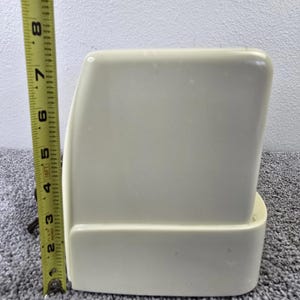 May include: A cream-coloured, rectangular object with rounded edges, approximately 15 cm tall. A yellow measuring tape is positioned next to the object, indicating its height. The object has a smooth, matte finish.