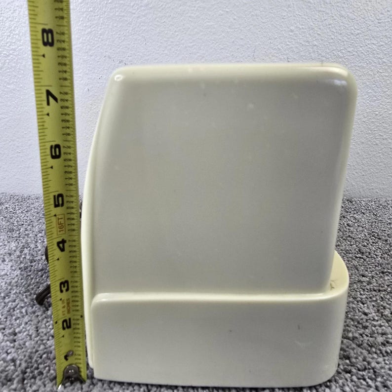 May include: A cream-coloured, rectangular object with rounded edges, approximately 15 cm tall. A yellow measuring tape is positioned next to the object, indicating its height. The object has a smooth, matte finish.