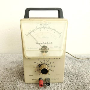 May include: A vintage beige Heathkit IM-38 multimeter with a black handle. The meter has a clear plastic cover over a gauge with a needle and a dial. It features a knob, input terminals, and the text "Heathkit".