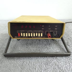 May include: A vintage Keithley 168 Autoranging DMM (Digital Multimeter) with a tan case and a dark face. The digital display shows "0255". It has various input jacks and function buttons. The device is supported by a dark metal frame.