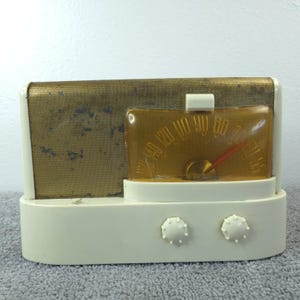May include: A vintage cream-coloured radio with a gold-toned speaker grill and a clear amber dial. The dial has a red needle and a numbered scale. Two white knobs are on the front.