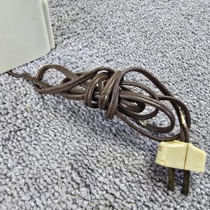 May include: A brown electrical cord with a cream-coloured plug. The cord is coiled and appears to be made of a flexible material. The plug has two prongs.