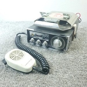 May include: A vintage Lafayette CB radio with a silver metal top, black face, and a white microphone. The radio has several knobs and a display. The microphone is connected to the radio with a black coiled cord. The radio is on a gray surface.