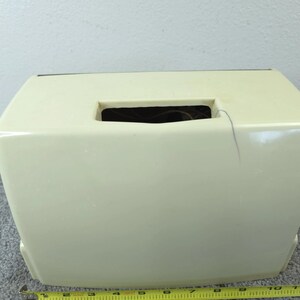 May include: A cream-coloured, rectangular tissue box with a rectangular opening on top. The tissue box is made of a smooth, glossy material. A yellow measuring tape is at the bottom of the image.
