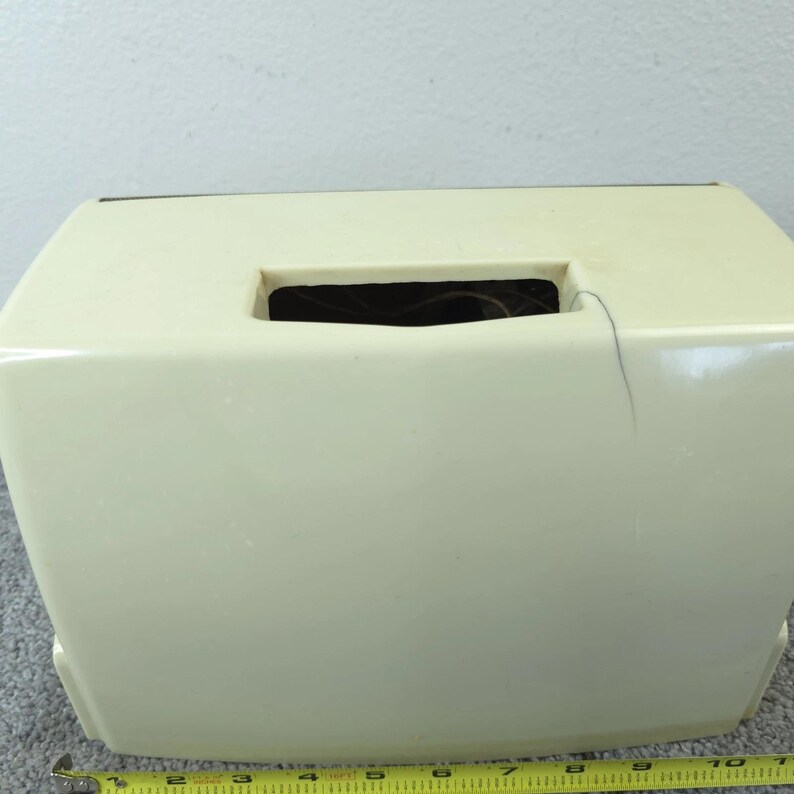May include: A cream-coloured, rectangular tissue box with a rectangular opening on top. The tissue box is made of a smooth, glossy material. A yellow measuring tape is at the bottom of the image.