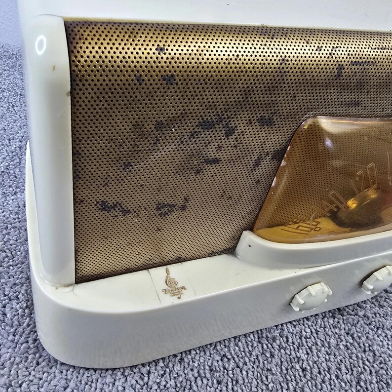 May include: Cream-coloured vintage radio with a gold-coloured perforated speaker cover. The radio has a clear, amber-coloured dial and two white control knobs. A small emblem is visible on the front. The radio is a retro design.