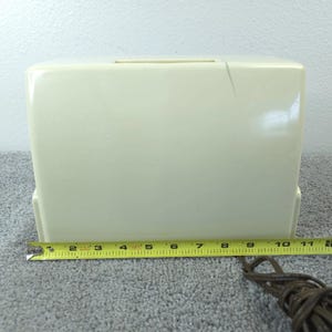 May include: Cream-coloured vintage sewing machine case. The case is rectangular with a handle slot on top. A yellow measuring tape shows the case is approximately 28 cm long. A brown electrical cord is visible.