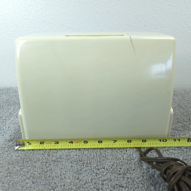 May include: Cream-coloured vintage sewing machine case. The case is rectangular with a handle slot on top. A yellow measuring tape shows the case is approximately 28 cm long. A brown electrical cord is visible.