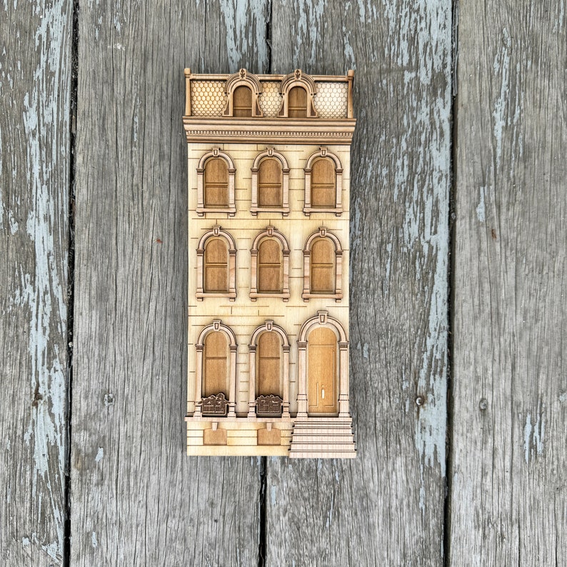May include: A wooden model of a three-story building with a detailed facade. The building has multiple windows and a door, all with intricate details. The model is painted in a light brown color.