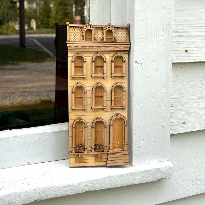 May include: A wooden model of a three-story building with a detailed facade. The building has a series of windows and a front door. The model is painted in a light wood tone.