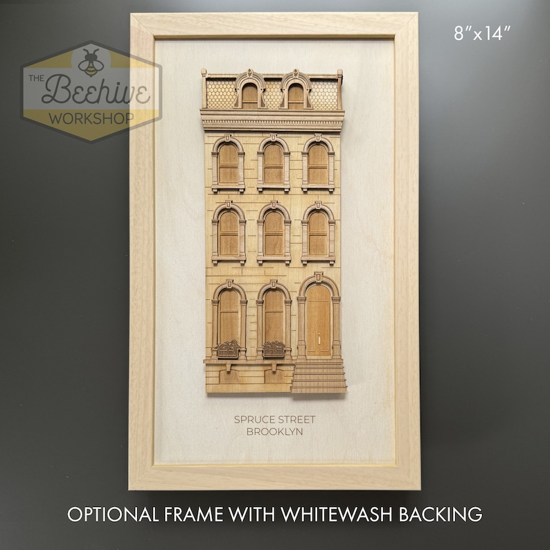 May include: A wooden model of a brownstone building with arched windows and a door. The model is framed in a light wood frame with a whitewash backing. The text "SPRUCE STREET BROOKLYN" is printed below the model. The frame measures 8 inches by 14 inches.