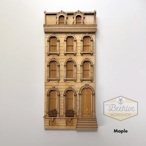 May include: A wooden cutout of a brown three-story building with arched windows and a door. The building has a detailed facade with decorative trim. The cutout is made of maple wood and is part of The Beehive Workshop.