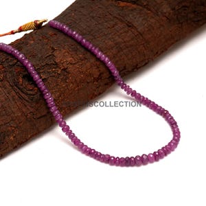 May include: A strand of small, faceted, deep pinkish-purple gemstone beads arranged in a circular loop. The beads are uniform in size and shape, featuring a reflective surface. A gold-toned metal clasp is visible at the end of the string.