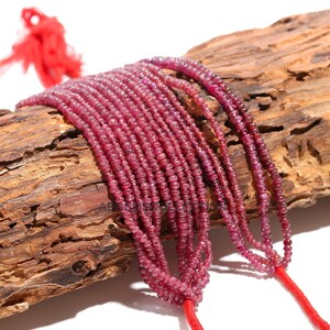 Op de afbeelding: Een streng roze robijn kralen, ongeveer 2 mm in diameter, geregen op een rood koord. De kralen zijn gefacetteerd en hebben een gladde, gepolijste afwerking. De streng wordt getoond op een stuk verweerd hout.