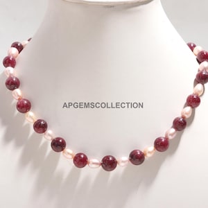 May include: A necklace featuring alternating dark red, round beads and light pink, oval-shaped pearls. The necklace is displayed on a white mannequin. The text "APGEMSCOLLLECTION" is visible on the mannequin.