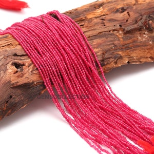 May include: A strand of bright pink gemstone beads, likely ruby, arranged on a piece of wood. The beads are small and uniform in size, creating a smooth and continuous line.