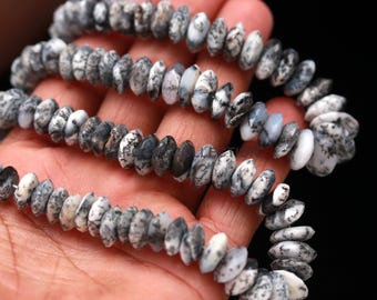 May include: A strand of gray and white gemstone beads. The beads are oval-shaped and have a speckled pattern. The beads are strung together, creating a necklace or bracelet. The beads are held in a hand.