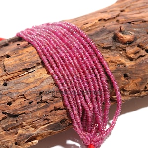 Op de afbeelding: Een streng roze robijn kralen, ongeveer 2 mm in diameter, geregen op een koord. De kralen zijn gefacetteerd en hebben een glanzende afwerking. De streng wordt getoond op een stuk hout.