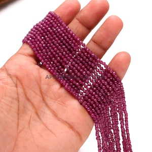 May include: Strands of small, faceted, deep red beads. The beads are held in a hand, showcasing their color and texture. The beads appear to be strung together, ready for jewelry making or other crafts.