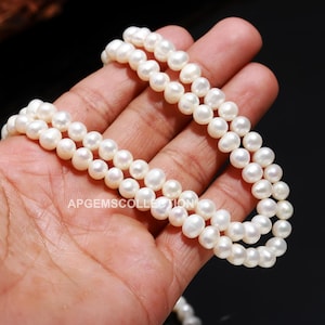 Könnte beinhalten: Eine doppelte Reihe weißer Süßwasserperlen. Die Perlen sind rund und glatt und auf einem weißen Faden aufgefädelt.