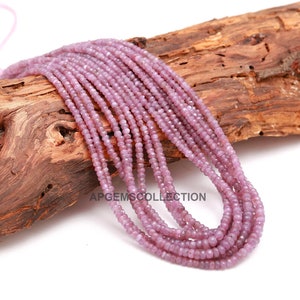 Könnte beinhalten: Eine Strähne rosafarbener Edelsteinperlen, etwa 2 mm groß, auf einem Stück Holz präsentiert. Die Perlen sind facettiert und haben eine leicht unregelmäßige Form.