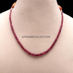Pode incluir: Um colar de fio único composto por pequenas contas de pedras preciosas facetadas em vermelho rosado profundo. O colar apresenta extremidades de cordão em tom laranja e exibe o texto APGEMSCOLLECTION sobre um fundo neutro.