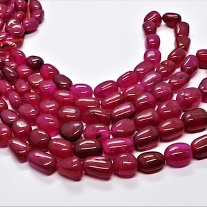 Könnte beinhalten: Eine Strähne aus rosafarbenen Achat-Edelsteinkugeln. Die Perlen sind glatt und poliert, mit einer leicht ovalen Form. Sie sind auf einer dünnen Schnur aufgefädelt und bereit für die Schmuckherstellung.