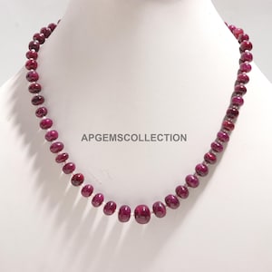 Op de afbeelding: Een ketting bestaande uit gepolijste, dieprode robijn kralen. De kralen zijn aan elkaar geregen, waardoor een doorlopende lus ontstaat. De ketting wordt getoond op een witte mannequin. De tekst "APGEMSCOLECTION" is zichtbaar.
