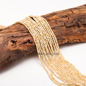 May include: A strand of white and cream colored round shell beads. The beads are approximately 4mm in diameter and have a slightly iridescent finish. The beads are strung on a clear thread and are displayed on a piece of wood.