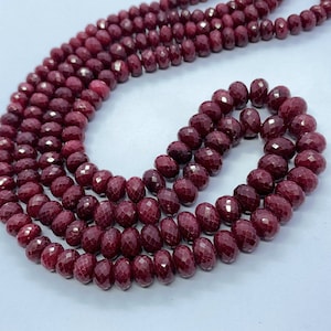 Può includere: Una collana di perle di rubino rosso scuro, sfaccettate e rotonde, infilate per formare una collana. Le perle sono disposte in un motivo sciolto e fluido, creando un aspetto visivamente accattivante ed elegante.