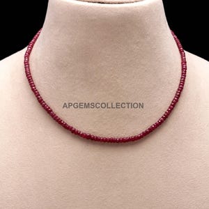 May include: A single strand necklace composed of small, faceted, deep red ruby beads. The necklace is displayed on a neutral-toned bust form. The text APGEMSCOLLECTION is printed in the center of the image, positioned just below the necklace strand.