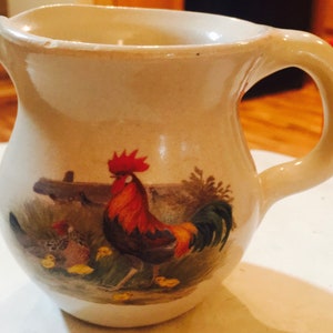 Puede incluir: Una pequeña jarra de cerámica de color crema con un gallo y gallinas pintadas en el lateral. El gallo es rojo, negro y amarillo, y las gallinas son marrones y blancas.