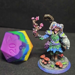 May include: A hand-painted miniature figure of a fantasy character with an owl, holding a flowering branch. Beside it is a large, multi-colored twenty-sided die. The figure is on a base with mushrooms.