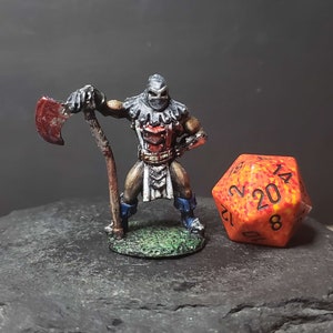 Puede incluir: Una figura en miniatura pintada de un guerrero con capucha sosteniendo un hacha con mango rojo. La figura está de pie sobre una base verde y está junto a un dado de 20 caras rojo y naranja.